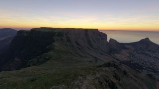 A flat-topped mountain rises above rugged green slopes and coastal neighborhoods, with the ocean beneath an orange horizon.