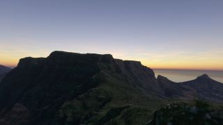 A flat-topped mountain rises above steep, shadowed slopes, with coastal buildings and the ocean beneath an orange horizon.
