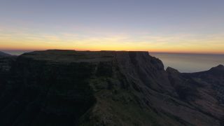 A flat-topped mountain rises above steep, shadowed cliffs, with the ocean stretching beneath an orange horizon.