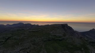 A rocky plateau with patches of green vegetation overlooks steep coastal cliffs and the ocean beneath an orange horizon.