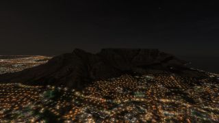 A flat-topped mountain rises above illuminated city streets at night, with a dark coastline extending to the right.