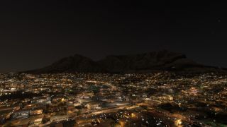 Illuminated streets and densely clustered buildings spread beneath a broad, flat-topped mountain under a starry night sky.