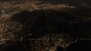 Dark mountain ridges rise above dense city lights at night, with illuminated roads tracing neighborhoods and the coastline.