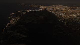 An aerial nighttime view shows dark mountain ridges surrounded by illuminated city streets, with coastal lights tracing the shoreline beside dark water.