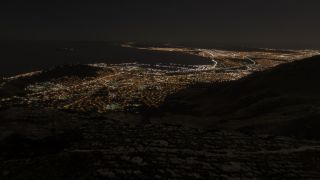 A coastal city glows at night around a broad bay, with illuminated roads tracing the shoreline and dark mountains in the foreground.