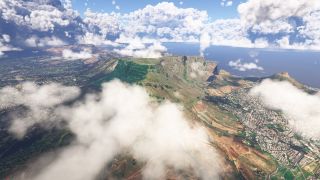 A flat-topped mountain rises above dense coastal neighborhoods, with scattered clouds obscuring its slopes and blue ocean stretching beyond.