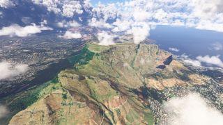 A flat-topped mountain rises above dense coastal neighborhoods, with steep rocky cliffs, winding hillside trails, and scattered clouds over the ocean.