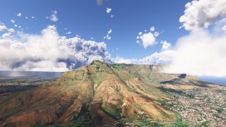 A flat-topped mountain rises above dense urban neighborhoods, with winding hillside trails, coastal water, and large clouds beneath a blue sky.
