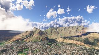A flat-topped mountain rises above dense urban neighborhoods, with steep rocky slopes beneath large cumulus clouds and blue sky.