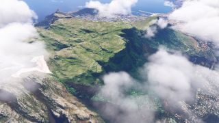 Scattered clouds partially obscure a broad, flat-topped mountain with steep green cliffs overlooking a coastal city and harbor.