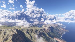 Steep coastal mountains rise beside a blue ocean, with scattered low clouds, towering cumulus formations, and urban development inland.