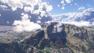 Low clouds partially obscure steep mountain cliffs above a coastal city, with winding hillside roads and harbor breakwaters visible below.