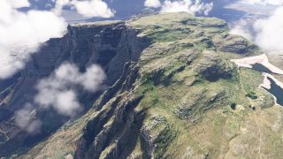 Scattered clouds partially obscure steep mountain cliffs, while grassy plateaus, winding paths, and two reservoirs remain visible from above.