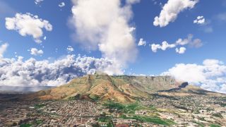 A flat-topped mountain rises above dense urban neighborhoods and green open spaces beneath large cumulus clouds in a blue sky.