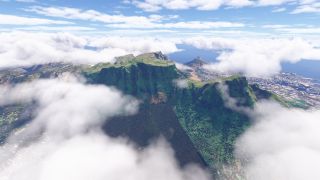 Low clouds surround a flat-topped mountain with steep green cliffs, while a coastal city and blue ocean extend beyond.