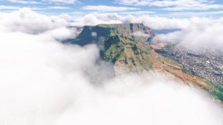 A flat-topped mountain rises through low clouds, with steep rocky slopes overlooking dense city blocks and winding roads.