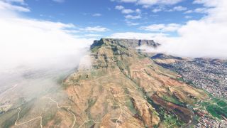 A flat-topped mountain rises above winding hillside trails and dense urban neighborhoods, with low clouds drifting across its rocky slopes.