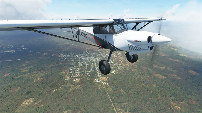 White XCub in flight over ground terrain with clouds in background.